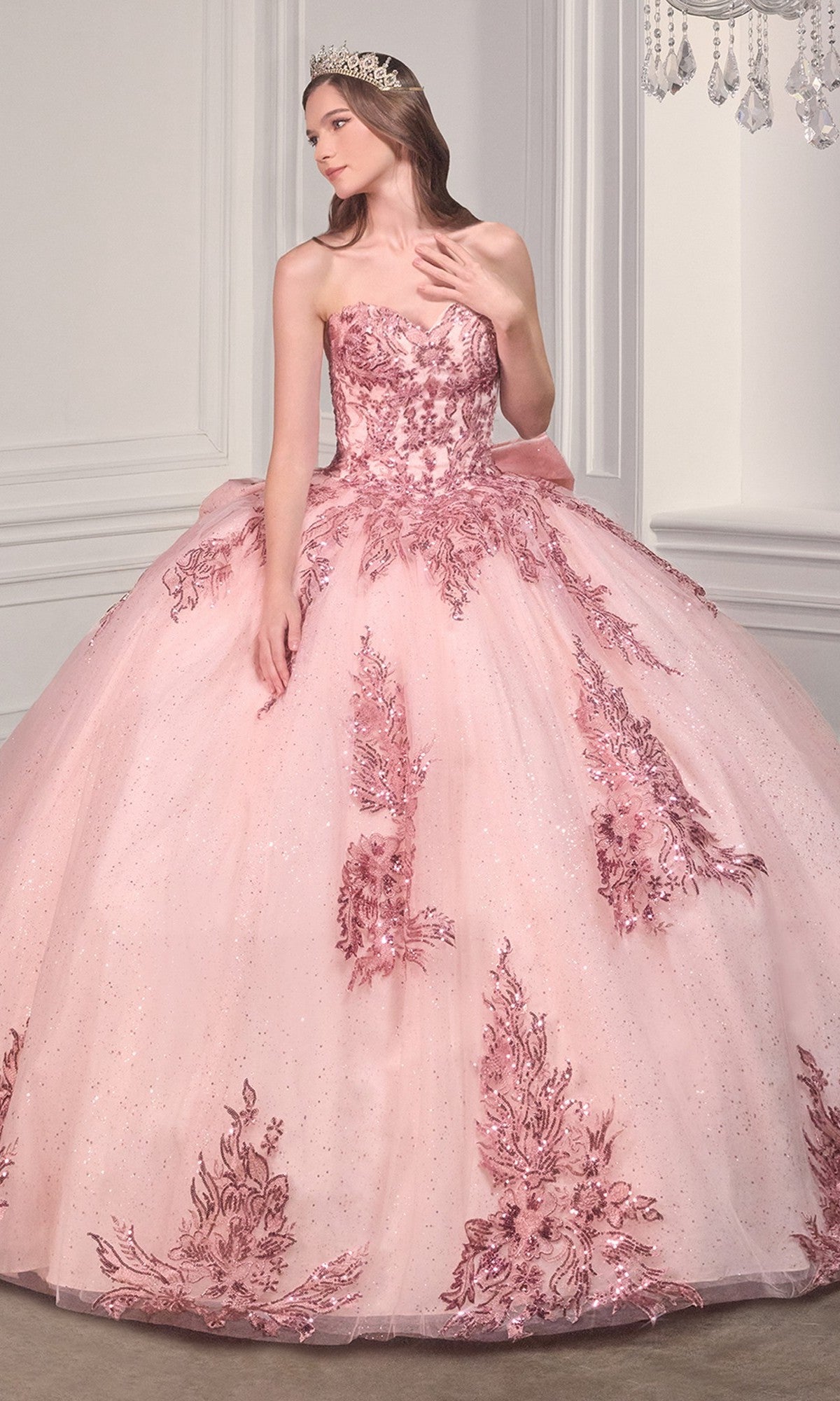 A woman stands in an elegant room wearing the Ladivine Strapless Quinceanera Dress 15715, a pink ball gown with floral embellishments, a bow detail, and a tiara against white paneled walls.